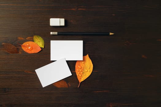 Photo of white business cards, pencil, eraser and bright autumn leaves on wooden background. Blank stationery template for placing your design.