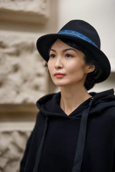 A girl of Asian appearance in a black blouse and hat, looking to the side against the background of a beige wall of a building on the street