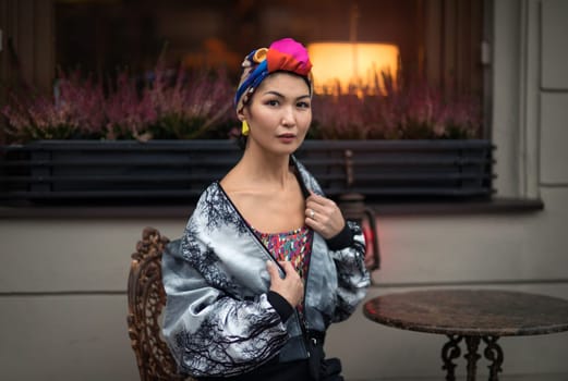 A girl of oriental nationality with a colored scarf on her head sitting on a chair at a small round table against the background of a pallet with heather