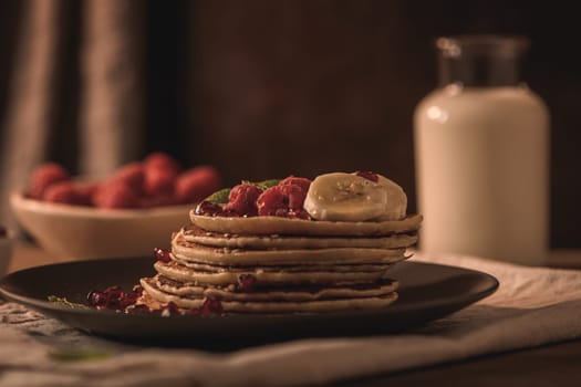 Pancakes with raspberries, banana slices, pomegranate seeds and honey on wooden vintage table.