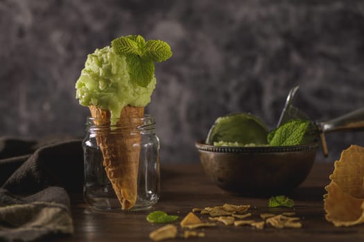 Pistachio ice cream in a dark rustic bowl and waffle cone on a brown wooden background.