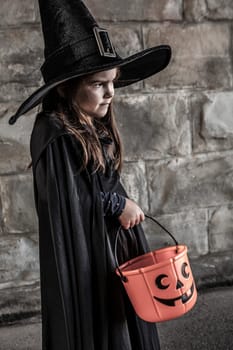 Young girl dressed in a witch costume with tall black hat and holding a pumpkin pail for halloween.