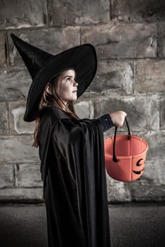 Young girl dressed in a witch costume with tall black hat and holding a pumpkin pail for halloween.
