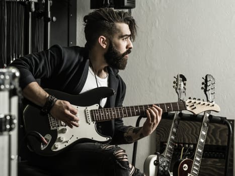 Photo of a man with beard sitting and playing his electric guitar in a recording studio..