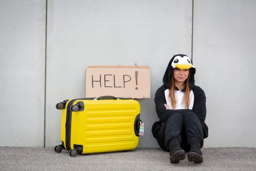 Young woman, with penguin costume and yellow suitcase, has to travel due to global warming. Symbolizes a sad penguin who has to leave his homeland because of global warming or climate change.