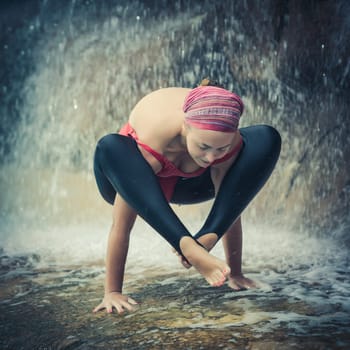 Woman practicing yoga near waterfall. Shoulder pressing posture. Bhujapidasana