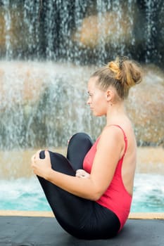 Woman practicing yoga near waterfall. Fetus Pose. Garbhasana