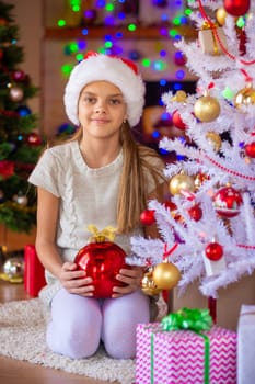 A girl with a red ball sits by the Christmas tree, in the background are blurry flashlights