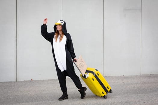 Young woman, with penguin costume and yellow suitcase, has to travel due to global warming. Symbolizes a sad penguin who has to leave his homeland because of global warming or climate change.
