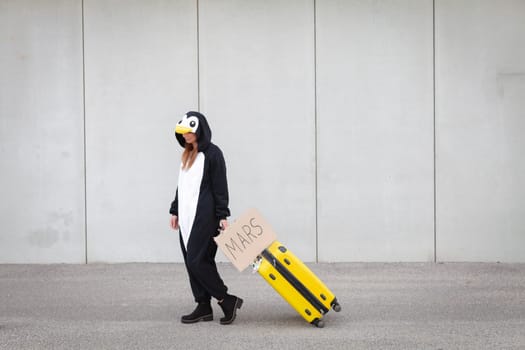 Young woman, with penguin costume and yellow suitcase, has to travel due to global warming. Symbolizes a sad penguin who has to leave his homeland because of global warming or climate change.