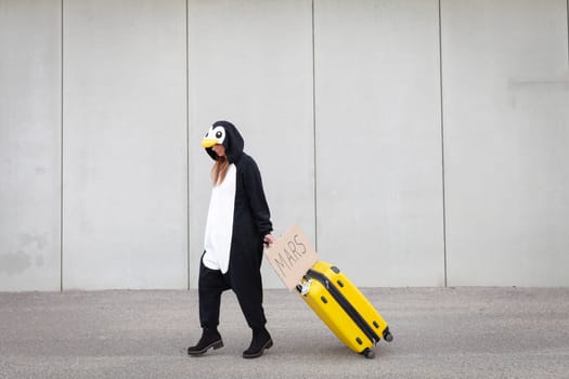Young woman, with penguin costume and yellow suitcase, has to travel due to global warming. Symbolizes a sad penguin who has to leave his homeland because of global warming or climate change.