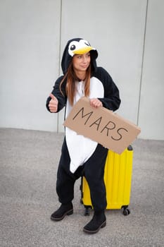 Young woman, with penguin costume and yellow suitcase, has to travel due to global warming. Symbolizes a sad penguin who has to leave his homeland because of global warming or climate change.