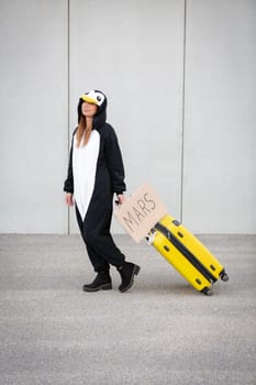 Young woman, with penguin costume and yellow suitcase, has to travel due to global warming. Symbolizes a sad penguin who has to leave his homeland because of global warming or climate change.