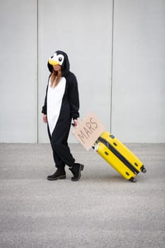 Young woman, with penguin costume and yellow suitcase, has to travel due to global warming. Symbolizes a sad penguin who has to leave his homeland because of global warming or climate change.