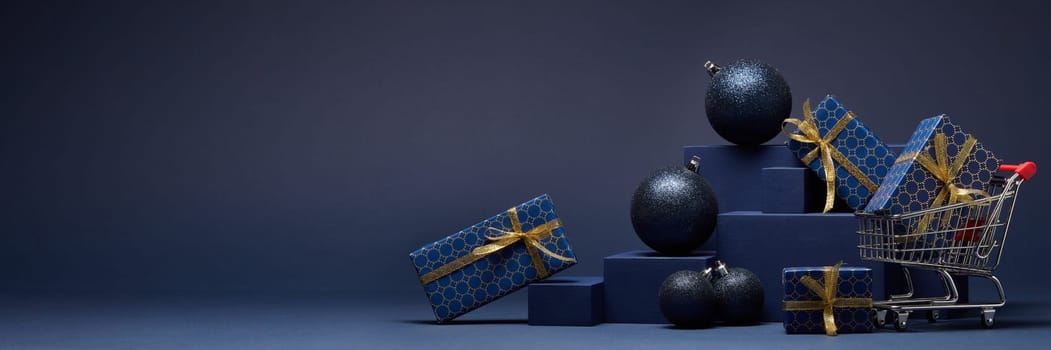 Shopping trolley, gift boxes and Christmas ornaments on dark blue background with copy space. Christmas and New Year shopping. Winter sale. For design of advertising, greeting card