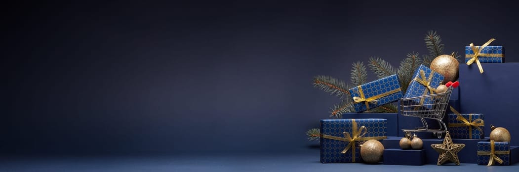Shopping trolley, gift boxes, fir branch and Christmas ornaments on dark blue background with copy space. Christmas and New Year shopping. Winter sale. For design of advertising, greeting card