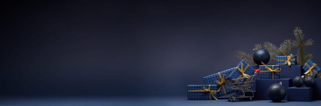 Shopping trolley, gift boxes, fir branch and Christmas ornaments on dark blue background with copy space. Christmas and New Year shopping. Winter sale. For design of advertising, greeting card