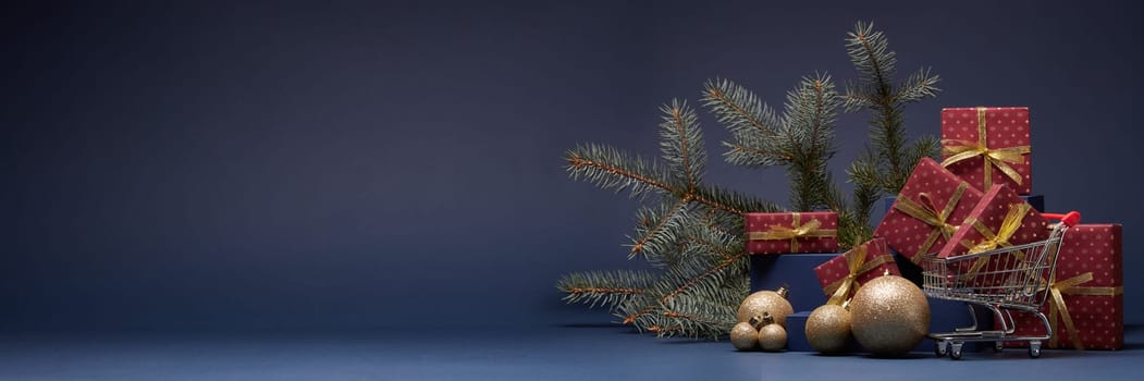 Shopping trolley, red gift boxes, fir branch and Christmas ornaments on dark blue background with copy space. Christmas and New Year shopping. Winter sale. For design of advertising, greeting card