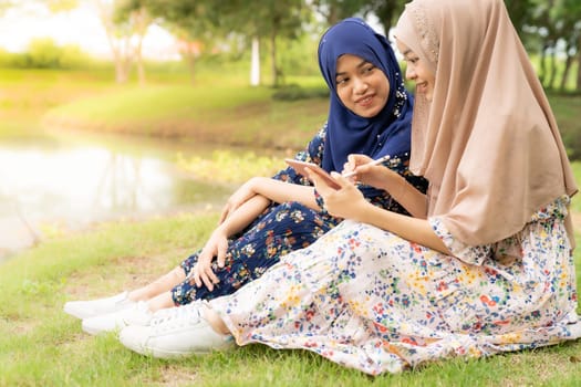 Young adult teenager Muslim Islamic Thai Asian women using smartphones and digital tablet in social media beside lake and green park in University college school.