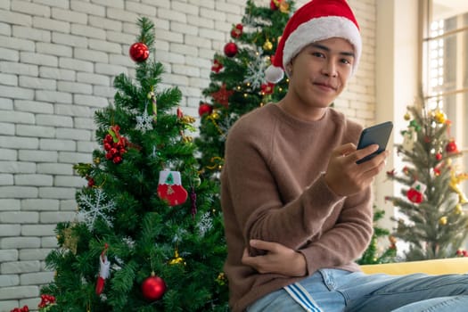 Portrait of good-looking smart Teenager young asian adult boy wearing brown sweater using his mobile phone for social media while waiting her girlfriens for Christmas celebration in living room with christmas decoration.
