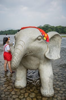 Guilin, China -  August 2019 : Chinese girl drying out her towel on top of the large marble elephant statue next to the landmark Elephant Trunk Hill Arch and the Li Lijiang River in Guilin, Guangxi Province