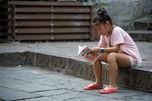 Feng Huang, China -  August 2019 : Young chinese girl sitting on a pavement and reading childrens book, Feng Huang Old Town