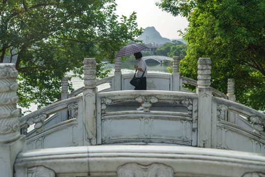 Guilin, China -  August 2019 : Chinese woman holding umbrella protecting her from the scorching summer heat and sun walking over one of the twin marble bridges on Shan Lake, Guangxi Province