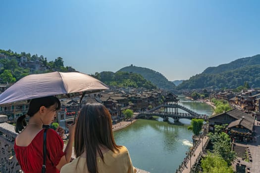 Feng Huang, China -  August 2019 : Women holding umbrella standing on the bridge and admiring the traditional old houses on the riverbanks of Tuo river in Feng huang Old Town