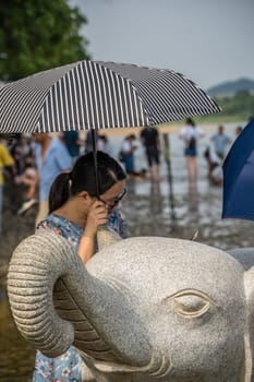 Guilin, China -  August 2019 : Woman holding umbrella to protect her from hot sun standing behind large marble elephant statuenear the landmark Elephant Trunk Hill Arch and the Li Lijiang River in Guilin, Guangxi Province