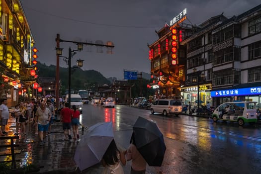 Wulingyuan, China - August 2019 : People walking in the rain with lights on buildings lit at night at a busy bustling high street in Wulingyuan town at dusk