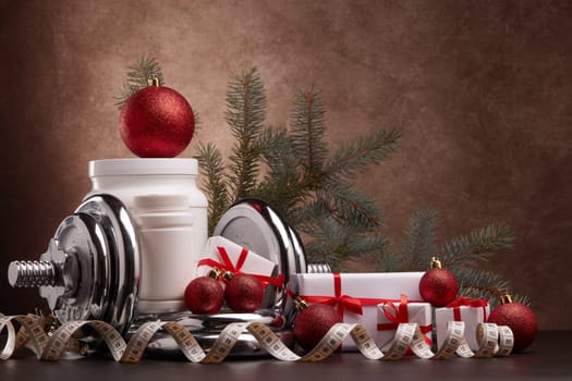 Sports nutrition (supplements), sports equipment, fir branch, gift boxes, Christmas ornaments on dark brown background. Fitness, sport, healthy lifestyle concept. New Year. Christmas.