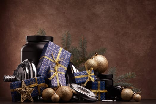 Sports nutrition (supplements), sports equipment, fir branch, gift boxes, Christmas ornaments on dark brown background with copy space. Fitness, sport, healthy lifestyle concept. New Year. Christmas.