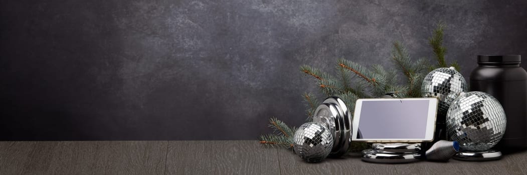 Sports nutrition, sports equipment, fir branch, Christmas ornament, blank computer tablet on dark grey background with copy space. Fitness, sport or healthy lifestyle concept. New Year and Christmas.
