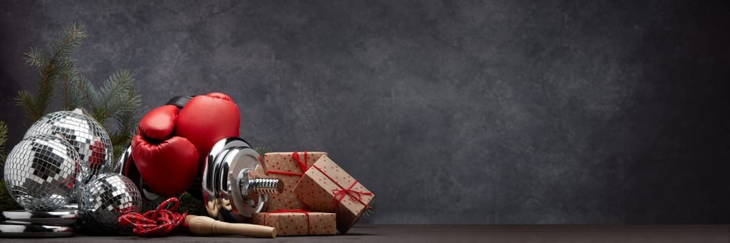 Sports equipment: dumbbell, boxing gloves, skipping rope, fir branch, Christmas ornament, gifts on dark grey background with copy space. Fitness, sport, healthy lifestyle concept. New Year. Christmas