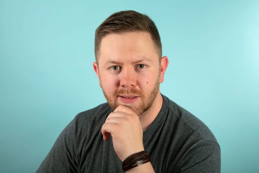 Bearded casual young man in a grey t-shirt looking pensively at the camera with his hand to his chin in a close up frontal studio portrait