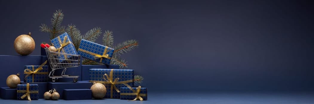 Shopping trolley, gift boxes, fir branch and Christmas ornaments on dark blue background with copy space. Christmas and New Year shopping. Winter sale. For design of advertising, greeting card