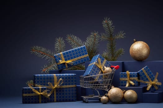 Shopping trolley, gift boxes, fir branch and Christmas ornaments on dark blue background with copy space. Christmas and New Year shopping. Winter sale. For design of advertising, greeting card