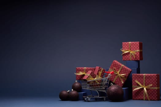 Shopping trolley, red gift boxes, fir branch and Christmas ornaments on dark blue background with copy space. Christmas and New Year shopping. Winter sale. For design of advertising, greeting card