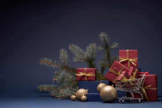 Shopping trolley, red gift boxes, fir branch and Christmas ornaments on dark blue background with copy space. Christmas and New Year shopping. Winter sale. For design of advertising, greeting card