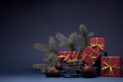 Shopping trolley, red gift boxes, fir branch and Christmas ornaments on dark blue background with copy space. Christmas and New Year shopping. Winter sale. For design of advertising, greeting card