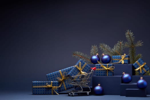 Shopping trolley, gift boxes, fir branch and Christmas ornaments on dark blue background with copy space. Christmas and New Year shopping. Winter sale. For design of advertising, greeting card
