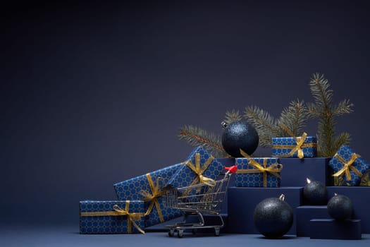 Shopping trolley, gift boxes, fir branch and Christmas ornaments on dark blue background with copy space. Christmas and New Year shopping. Winter sale. For design of advertising, greeting card