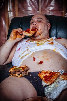 Fat couch potato eating a huge hamburger and watching television.  Harsh lighting from the television illuminates the dark room.