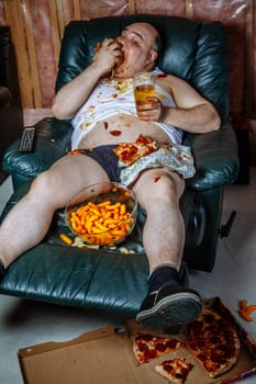 Fat couch potato eating a huge hamburger and watching television.  Harsh lighting from the television illuminates the dark room.
