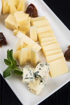 Cheese plate on a dark background table. Many kinds of cheese with sauce and greens on a white plate, closeup shot
