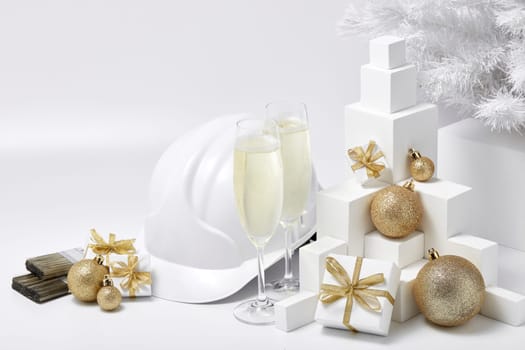 Construction hard hat, white fir tree, two glasses with champagne, gift boxes and Christmas ornaments on a white background. New Year and Christmas construction background