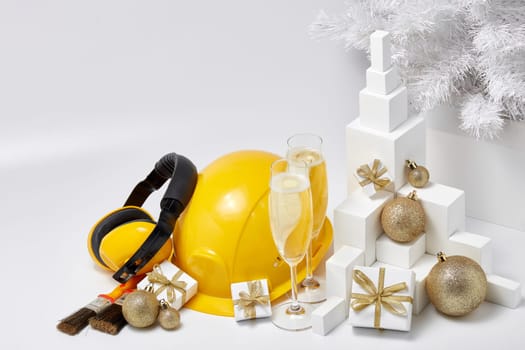Construction protective earphones and hard hat, fir tree, flutes with champagne, gift boxes and Christmas ornament on a white background. New Year and Christmas construction