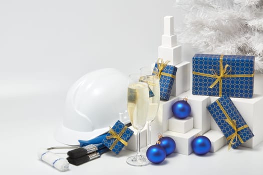 Construction hard hat, white fir tree, two glasses with champagne, gift boxes and Christmas ornaments on a white background. New Year and Christmas construction background