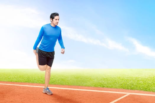 Asian runner man stretching legs before start the run on the running track with a blue sky background