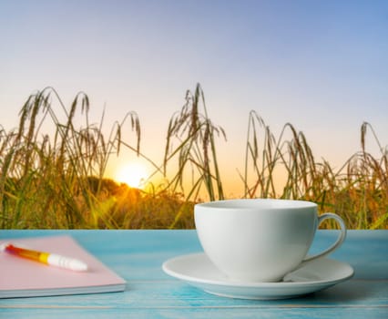 A cup of coffee and notebook with blurred rice field rural with colorful of sky in morning light, Relaxation and remember concept